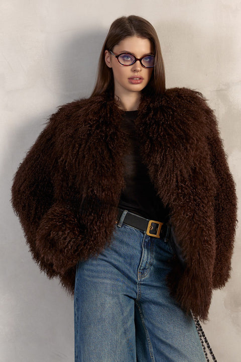 A woman wears a brown Mongolian coat handcrafted from the thickest, softest genuine Mongolian fur, featuring a dramatic collar, long sleeves, and elegant snap and hook closures.
