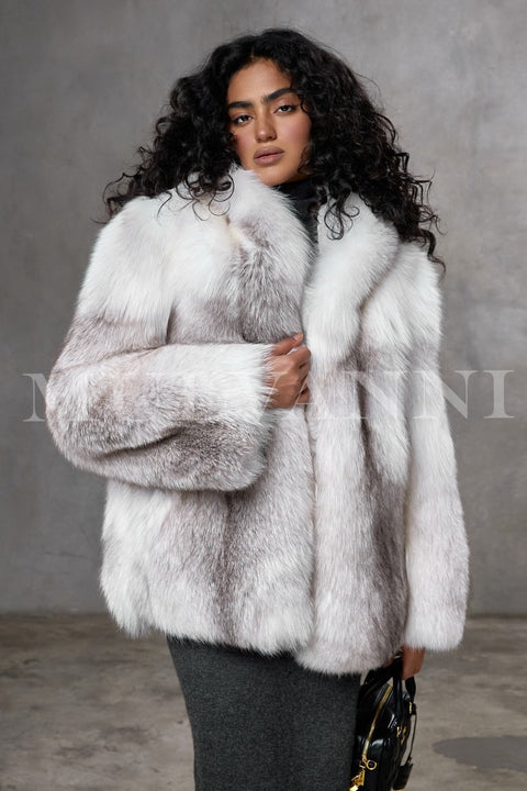 A woman wears a meticulously handcrafted full-pelt Arctic fox fur coat, featuring exceptional softness, volume, and a loose fit with a plush hood that envelops her in luxurious warmth and elegant style.